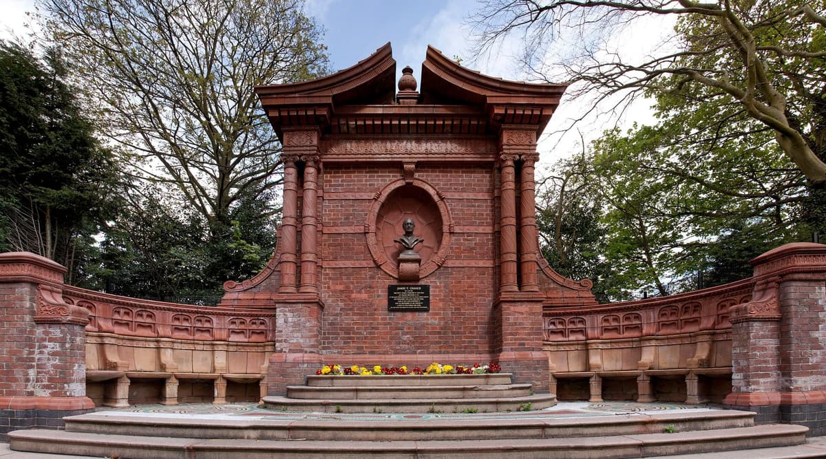 Red brick and terracotta memorial with a bust in a round-headed niche and curved seats on either side, in a park.