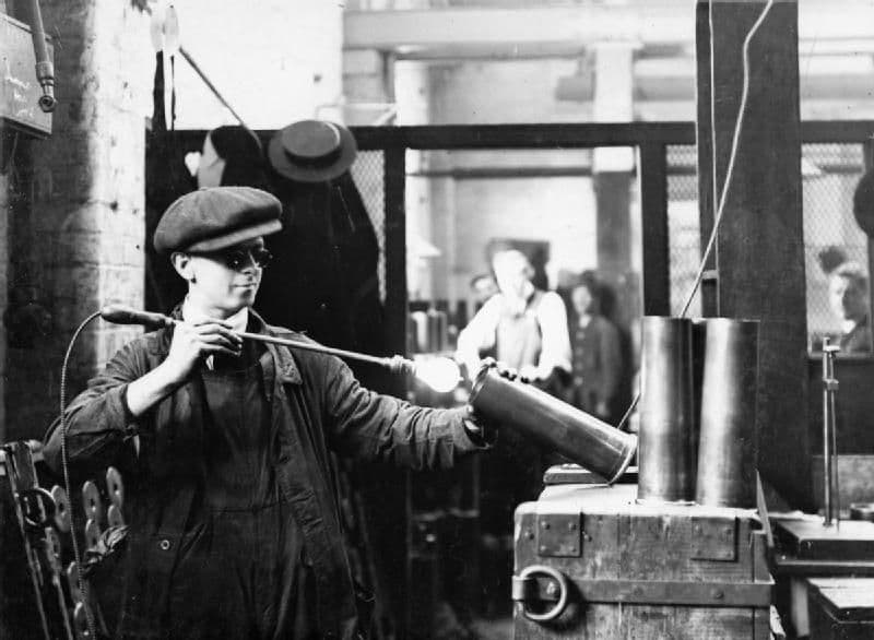A worker looking into a brass shell case lit from inside by an electric lamp.
