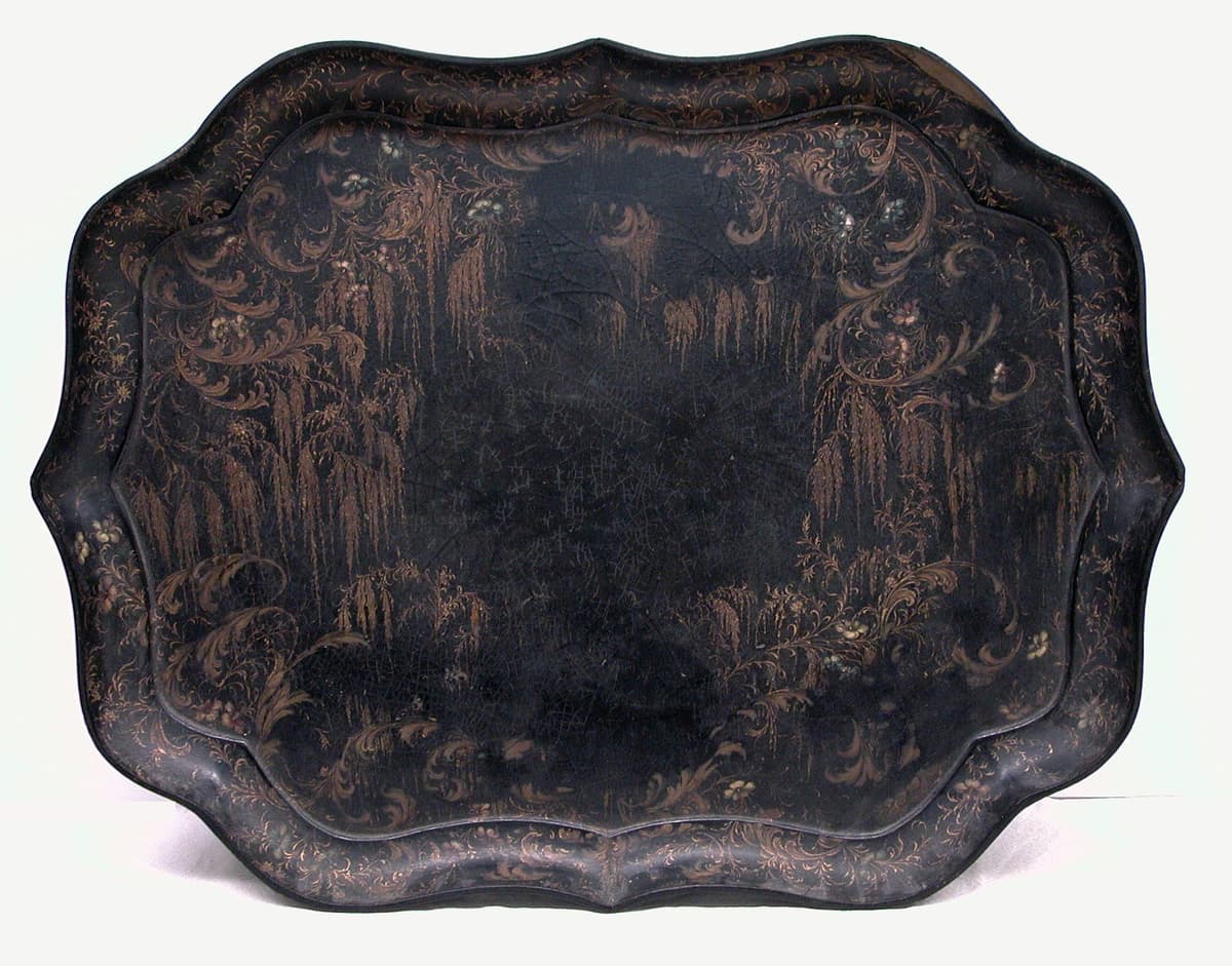 A black papier-mâché tray with a shaped edge, painted with gold scrolls, weeping willows and small flowers.