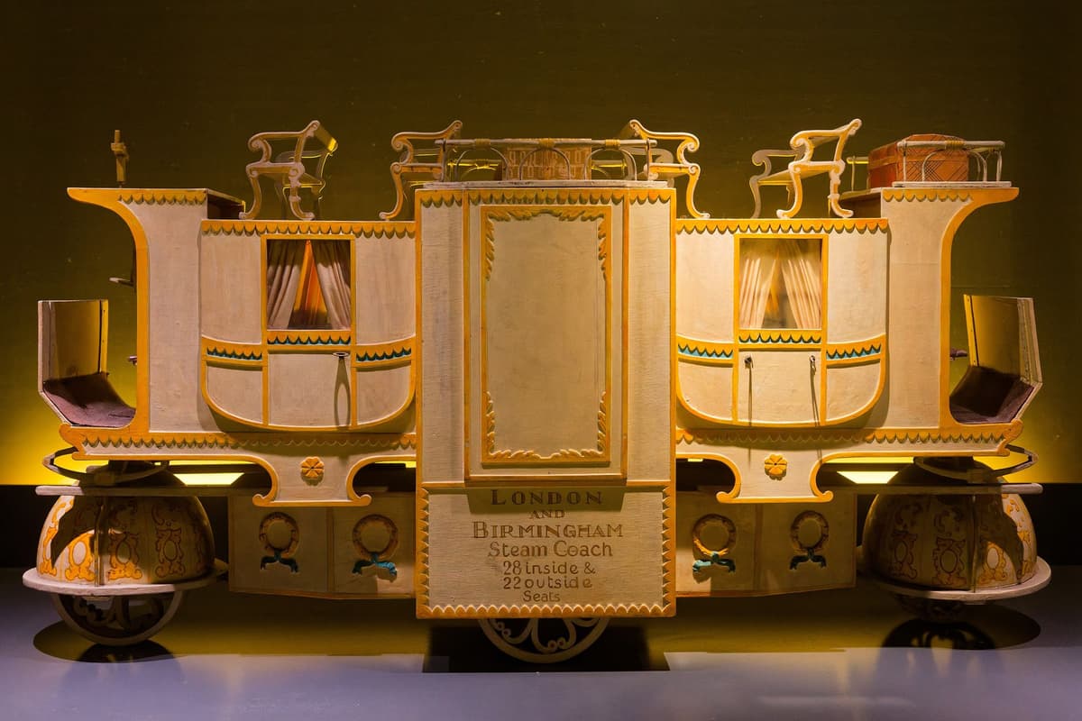 A cream and gold model of a long steam coach with two enclosed compartments, seats on the roof and a panel reading ‘London and Birmingham Steam Coach, 28 inside & 22 outside seats’.