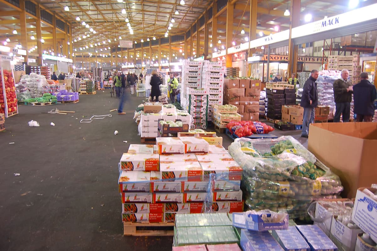 A large covered market hall with stacked produce and traders before dawn.