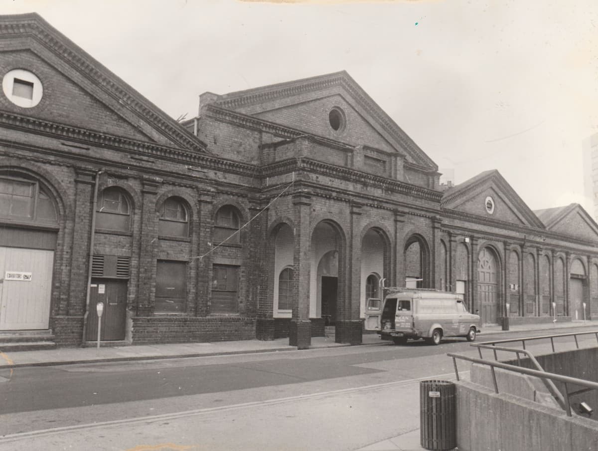 Black and white photograph of a brick building front with a pediment and round window above four open arches.