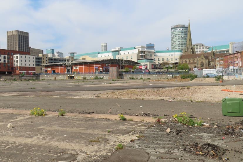 A wide cleared city-centre site behind fencing, with tower blocks beyond.