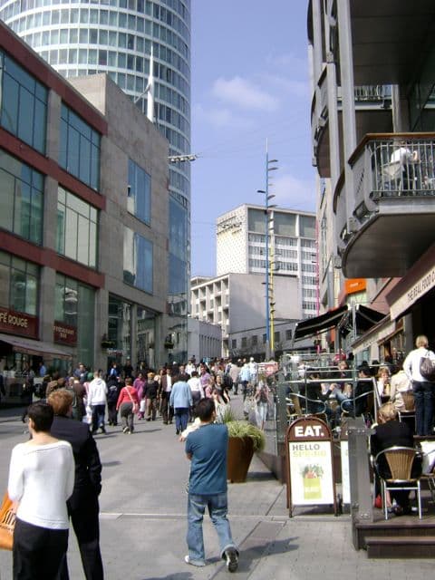 A wide pedestrian street climbing between modern shopping-centre buildings towards a cylindrical tower.