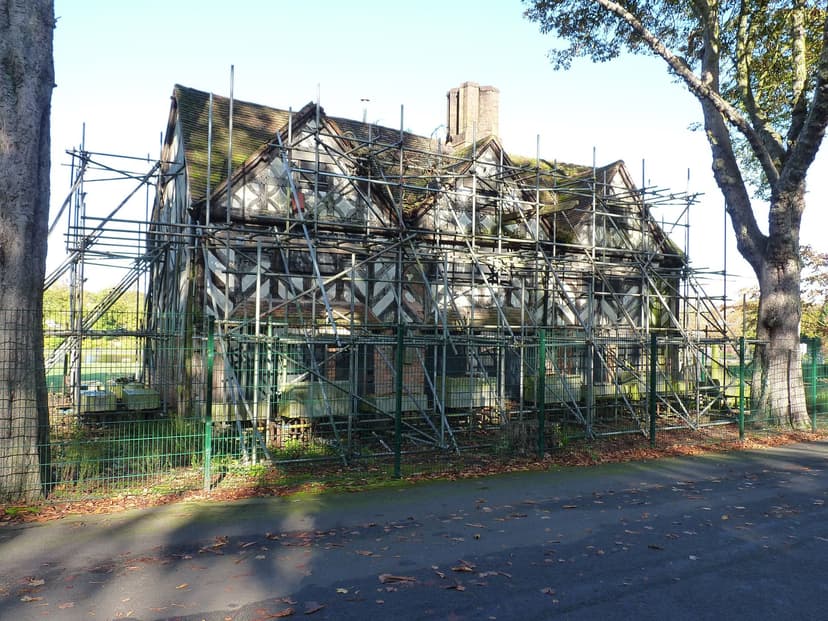 A timber-framed house with three gables wrapped in scaffolding in a park.