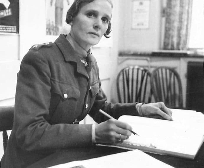 A woman in ATS uniform sits at a desk writing in a ledger with a pencil, looking at the camera; posters and wooden chairs are behind her.