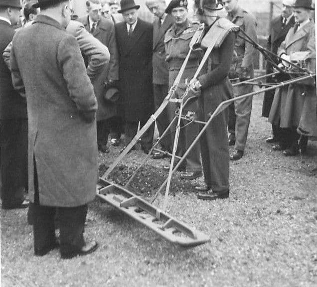 Men in overcoats and trilbies and officers in berets watch a man push a long wheeled frame, a Widesweep mine detector, across gravel outside a works.