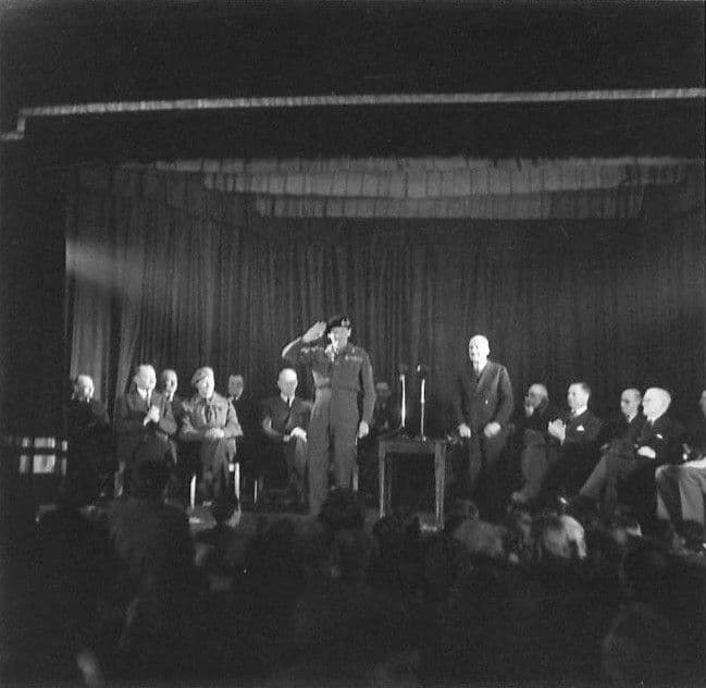 On a curtained stage lit by a spotlight, General Montgomery salutes beside a microphone while seated officials watch; an audience fills the dark foreground.