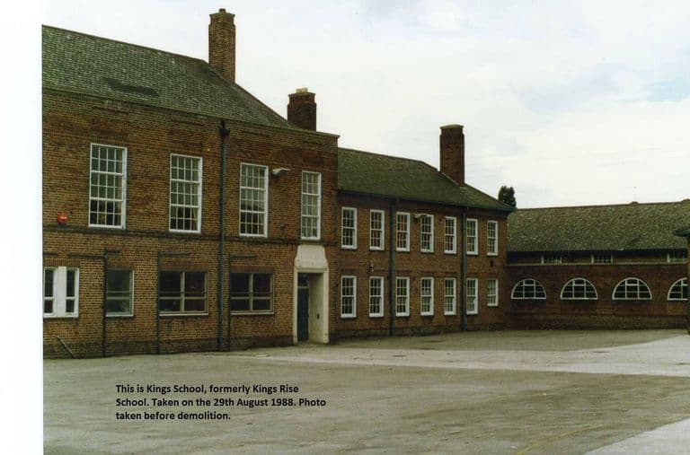 A red-brick two-storey school with sash windows, tall chimneys and a range of half-moon windows, beside an empty playground