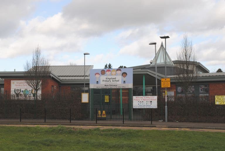 Single-storey brick school buildings with a modern glazed pyramid roof, railings and a school sign, grass in front