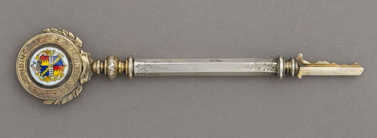 Ornate silver ceremonial key with an enamelled Birmingham coat of arms in a laurel-wreath bow, photographed on a grey background