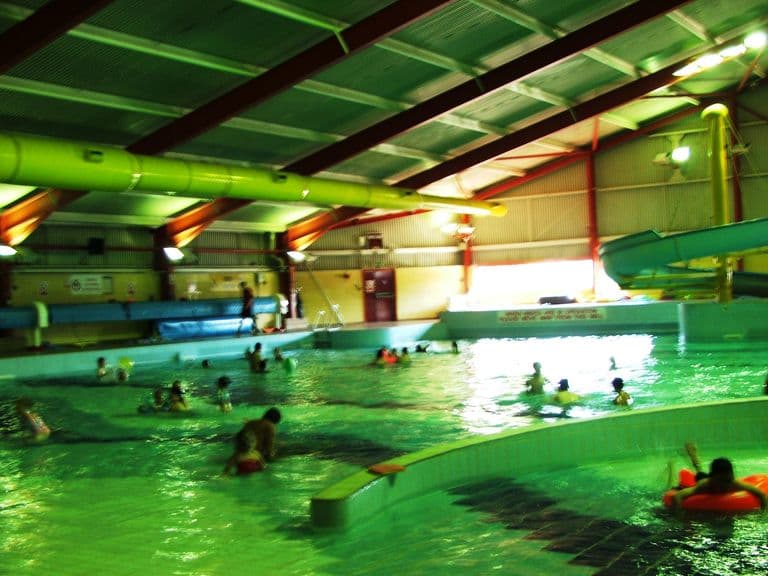 Indoor leisure pool with green-lit water, a spiral water flume, swimmers and a steel roof, at Kingstanding Leisure Centre