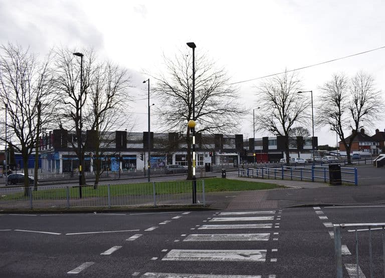 Row of flat-roofed 1960s precinct shops behind bare trees across the Circle from Kettlehouse Road, with a pedestrian crossing in front