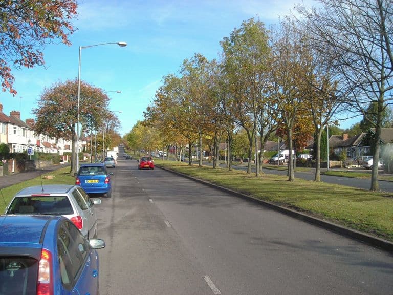 Wide tree-lined Kingstanding Road in autumn with a central grass verge, parked cars and inter-war semi-detached houses on both sides