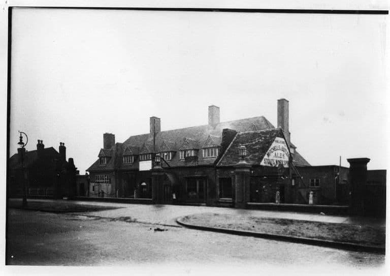 A large inter-war pub with dormer windows and tall chimneys, a gable painted Mitchells & Butlers Ales, Stouts, Wines & Spirits, behind brick gate piers with lamps