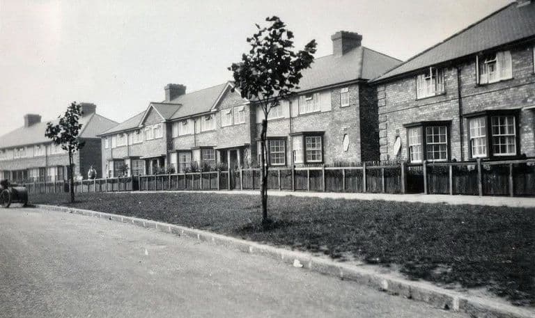 A row of new brick council houses with wooden paling fences, a wide grass verge and a freshly planted young tree; a motorcycle and sidecar at the left
