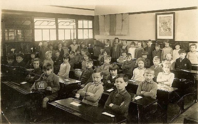 Black-and-white class photograph of about forty children sitting at wooden double desks in a classroom, a boy in front holding a slate marked VIII