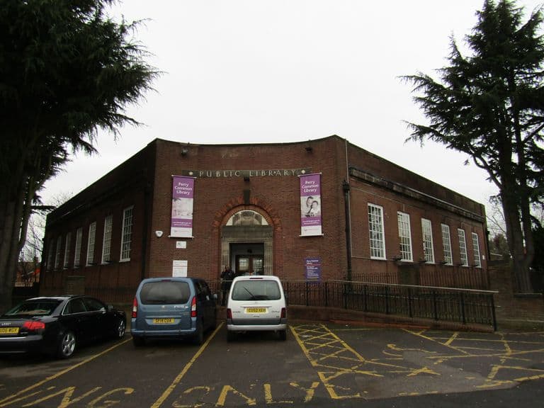 Single-storey brick library with a curved corner, 'PUBLIC LIBRARY' lettering, an arched stone entrance and purple banners, cars parked in front