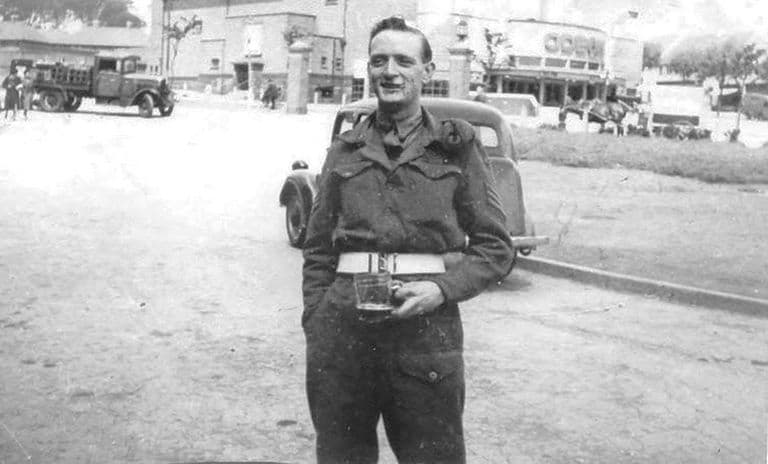 A smiling soldier in battledress and white belt holding a glass of beer, with a lorry, a car and the Odeon cinema behind him