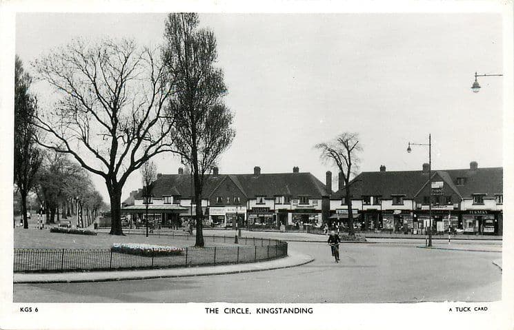 1950: Black-and-white postcard of the Circle, Kingstanding, around 1950: a parade of shops behind the railed garden in the middle of the Circle, with a cyclist on the road