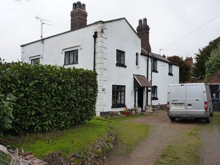 White-painted two-storey cottage row with a big brick chimney stack and small windows, a van on the gravel drive and hedge in front