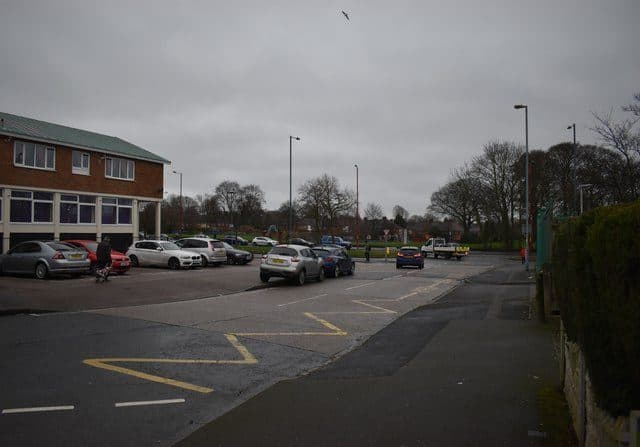 A wide road junction on Kingstanding Road with parked cars and the open grass of Wheeldon Recreation Ground beyond
