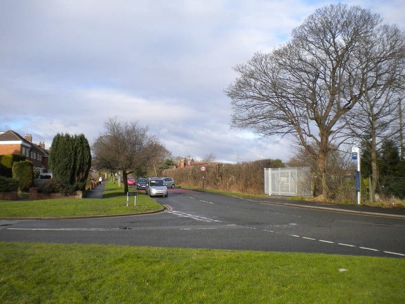 Road junction with trees and a fenced compound on the right, at the end of Doe Bank Lane