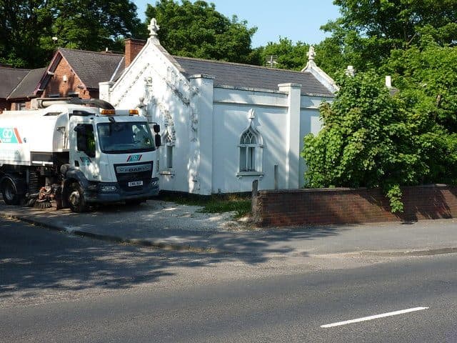 A small white single-storey Gothick lodge with pointed windows and a gabled porch, behind a low wall, with a lorry parked outside