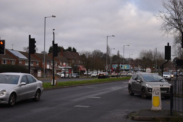 A dual carriageway at traffic lights, with cars and a row of shops and houses beyond