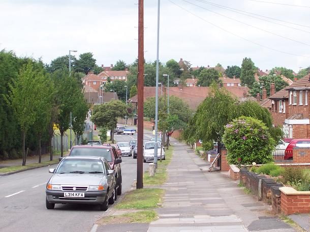 A suburban road with parked cars, a pavement and trees, sloping down towards houses