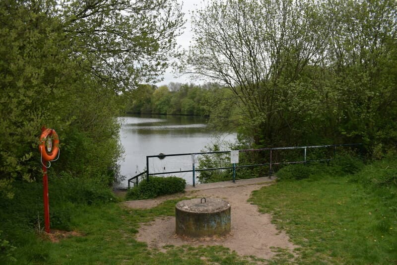 The lake at Queslett Nature Reserve, seen between spring trees from a sandy clearing with a lifebuoy post and a low railing