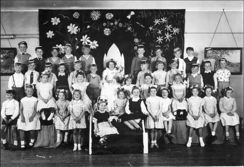 An infant-school May Day group in a hall: a May Queen in a white dress and veil sits on a stepped throne in front of a large painted backdrop of daisies, with about forty children in party dresses, flower garlands and paper hats around her.
