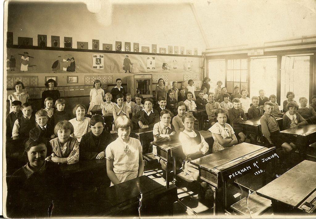 A sepia classroom photograph: about forty-five children at wooden double desks turn to face the camera, with children’s drawings and posters pinned along the wall and tall windows on the right. The front desk is chalked “Peckham Rd Junior 1935”.