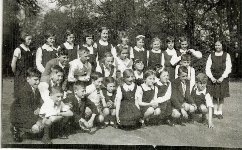 A class of about thirty boys and girls posed outdoors in front of trees, the girls standing behind in white blouses and dark pinafores and the boys crouching at the front.