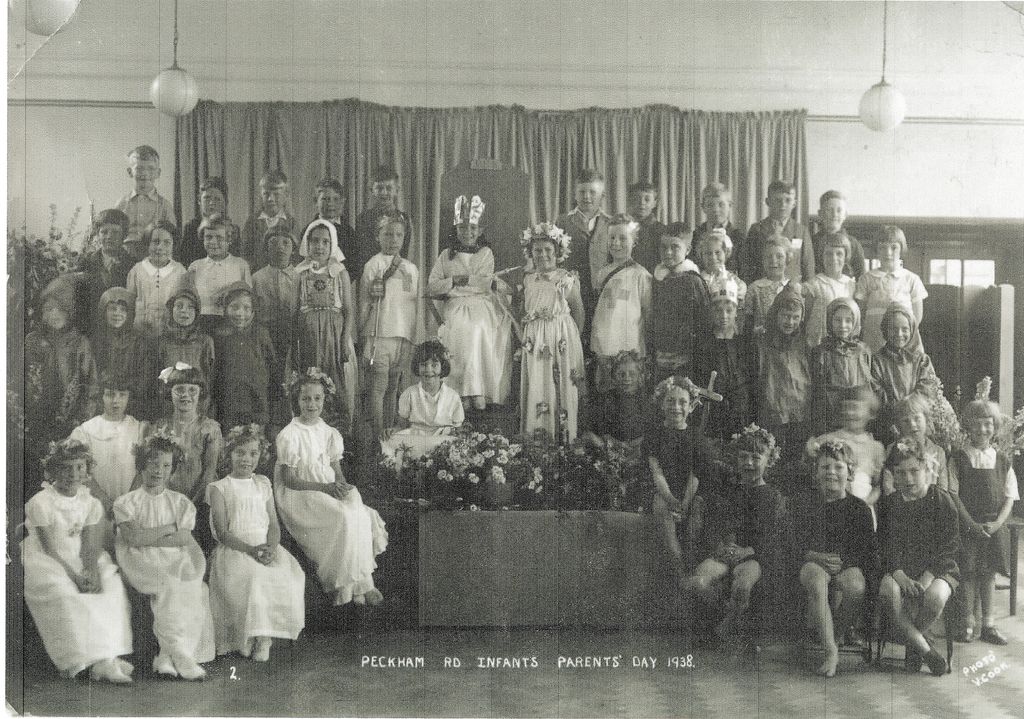 About fifty infants in costume on a stage in front of a curtain: a child in a crown sits on a throne among girls in white dresses with flower garlands, boys in tabards, and a bank of flowers at the front. The print is titled “Peckham Rd Infants Parents’ Day 1938”.