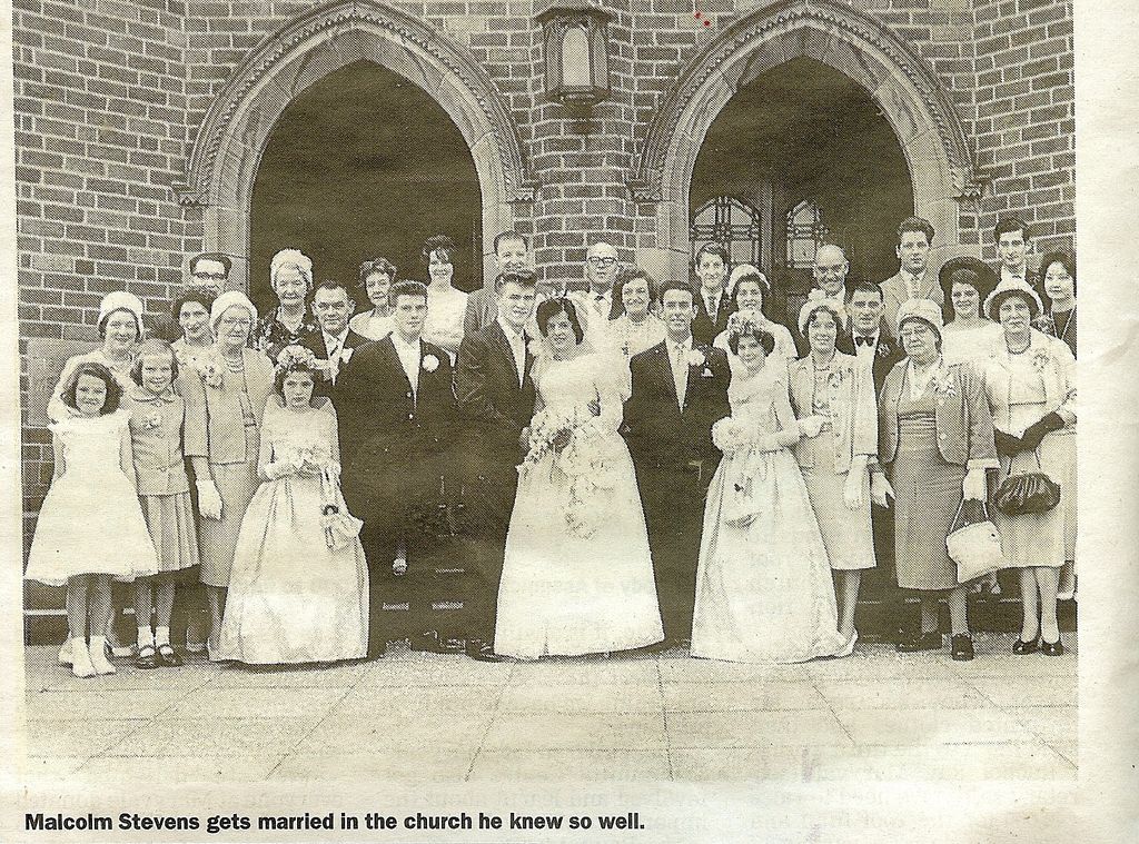 A newspaper photograph of a wedding party of about forty people in front of the church’s two pointed-arch brick doorways: the bride and groom in the centre with bridesmaids and flower girls in long dresses, guests in hats and suits. The printed caption reads “Malcolm Stevens gets married in the church he knew so well.”