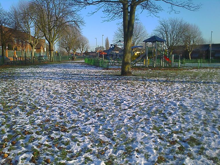 A park lawn dusted with snow and fallen leaves under bare trees, with a fenced children’s play area with a slide and climbing frame on the right and houses behind.
