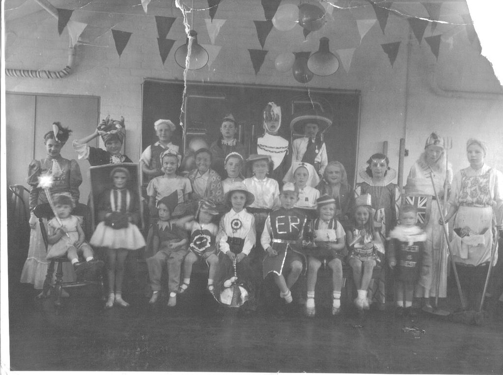 Twenty-four children in fancy dress – a Queen’s coach guard with “ER” on his tabard, a knight, a clown, fairies, a nurse, a girl dressed as a doll in a box – pose in a room hung with bunting and balloons. The print is creased and torn at the top right.