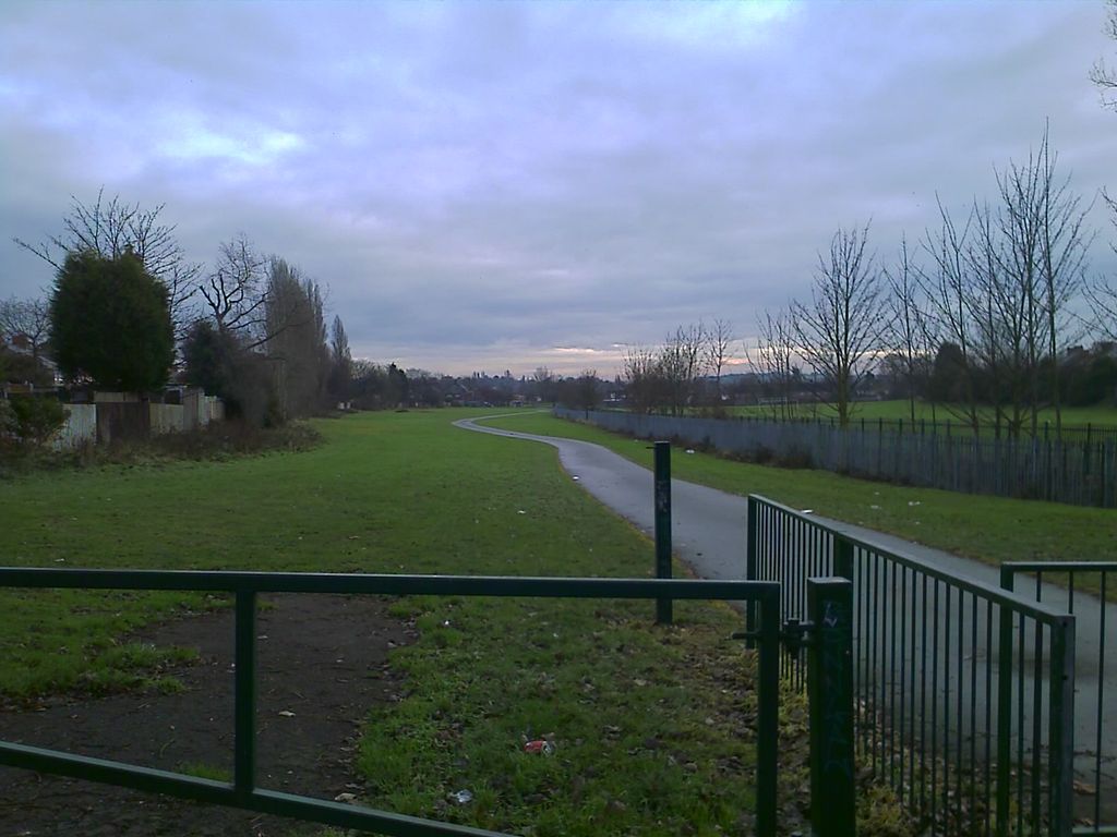 A long strip of open grass with a winding tarmac path, bounded by railings and a palisade fence and lined with young trees, seen past a green metal gate under a cloudy sky.