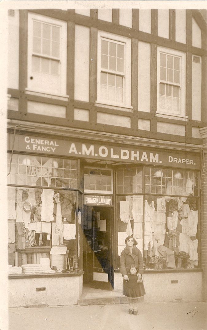 A mock-Tudor shop front with the fascia “General & Fancy A. M. Oldham Draper” and windows full of clothes and linens; a woman stands in the doorway with a small girl in front of her. A sign over the door reads “Agent for Paragon Laundry”.