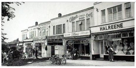 A parade of shops with flats above: “Allen’s Cycle Radio” with its sign on the upper wall, a shop lettered “Halreene”, bicycles propped at the kerb, shoppers on the pavement and a horse and cart at the left.
