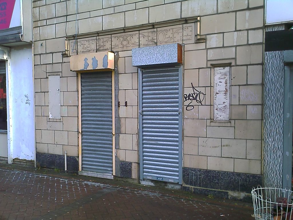 Two narrow doorways closed with metal roller shutters in a pale faience-tiled shop front, beneath a moulded frieze of stylised flowers; graffiti on the wall and a block-paved pavement in front.
