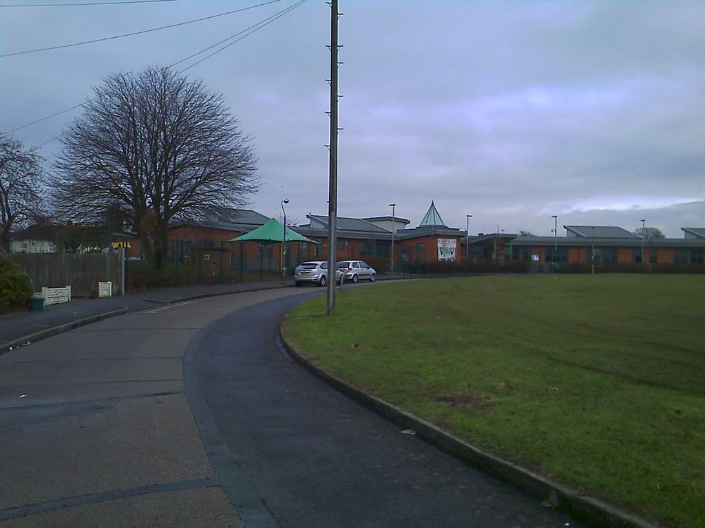 Low modern school buildings with a pointed turret roof and a green canopy behind railings, seen across a curving road and a large grassed area under a grey sky.