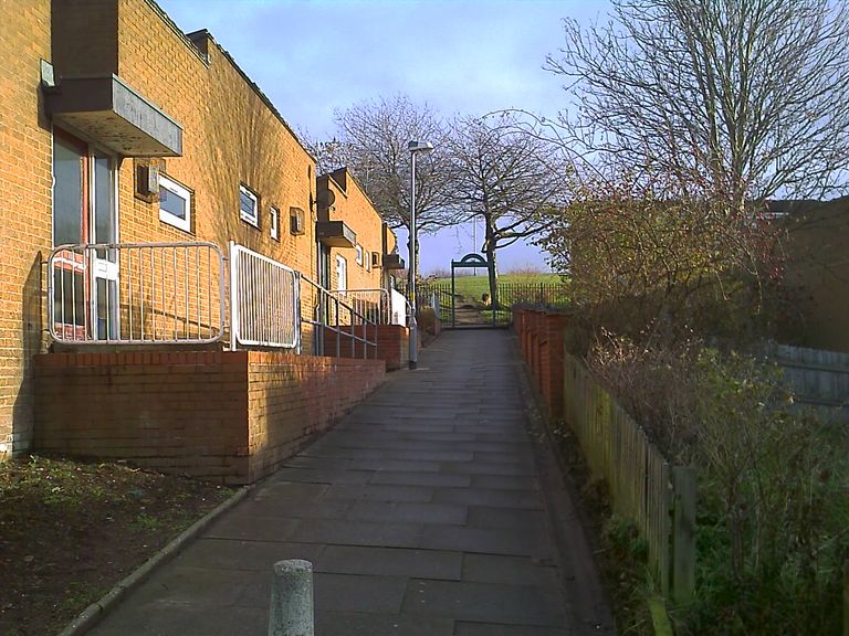 A paved footpath runs between a terrace of brick bungalows with small balconies and a wooden fence towards a gate in green railings, beyond which a grassy slope rises to the skyline.
