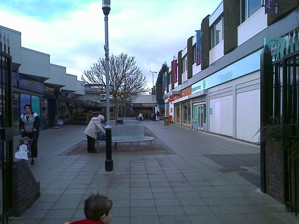 A paved pedestrian shopping precinct with a lamp post, a circular bench round a young tree and two-storey shop units on both sides; shoppers walk through, and a child’s head is in the foreground.