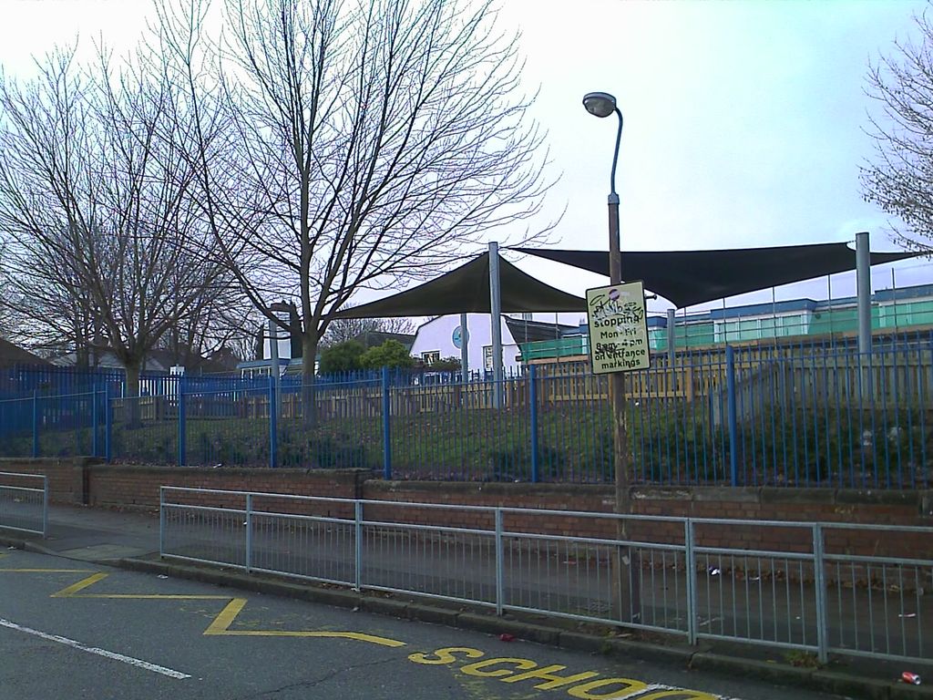 Modern school buildings with shade sails over the playground, seen behind blue railings and a low brick wall from Cranbourne Road; bare trees, a lamp post with a no-stopping sign and “SCHOOL” road markings in front.
