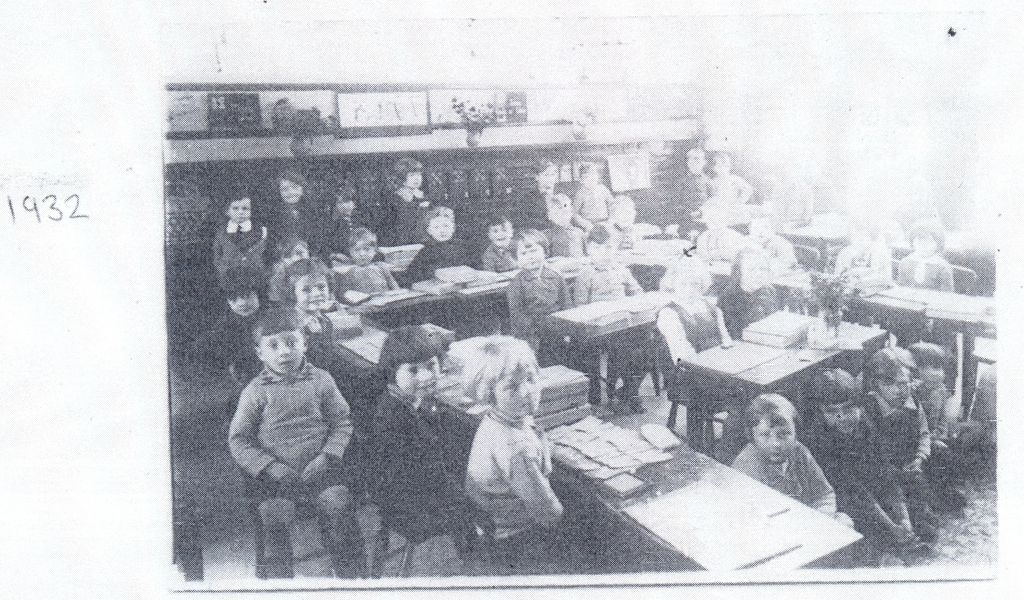 A faded photocopy of a classroom photograph: rows of small children at wooden desks with slates and papers, pictures along the wall and a vase of flowers; “1932” is handwritten beside it.