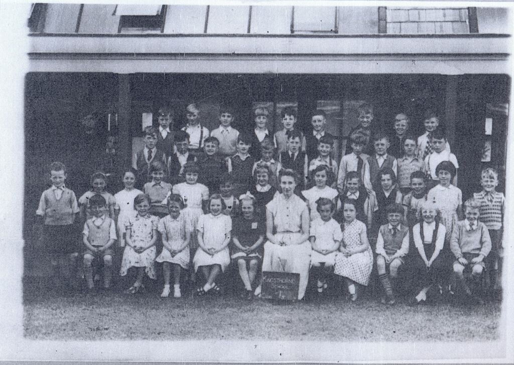 A class of about forty-five children in rows on grass in front of a verandah, with a woman teacher seated in the centre; a slate at her feet reads “Kingsthorne School”.