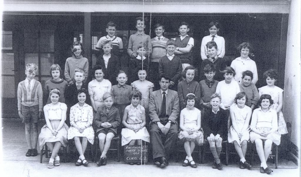 A class of about thirty-five children in four rows with a young male teacher seated in the centre, under a verandah; a slate at the front reads “Kingsthorne Junior School 1962 Class 4.2”.
