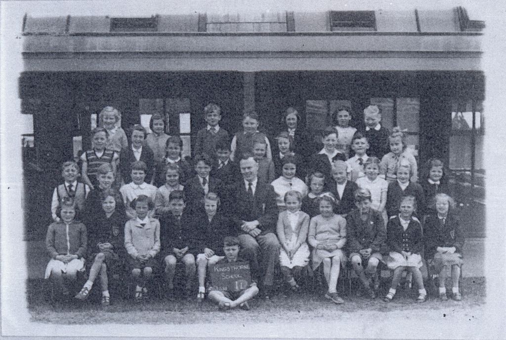 A class of about forty children in four rows with a male teacher seated in the centre, in front of a verandah and windows; a boy at the front holds a slate reading “Kingsthorne School”.